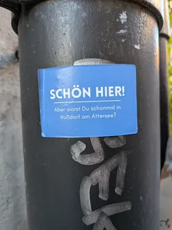 Street sticker A rectangular sticker with slightly rounded corners, featuring a solid blue background. The text is white and in German. The top line, 'SCHÖN HIER!', is larger and bold, followed by a thin white horizontal line. Below the line, the text continues in a smaller font: 'Aber warst Du schonmal in Nußdorf am Attersee?'. The sticker is applied to a dark, textured pole, with some faint graffiti visible beneath it.