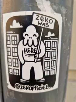 Street sticker Black and white sticker featuring a cartoon character holding a sign that says "ZeKo was here". The character is standing in front of buildings. The sticker includes the artist's Instagram handle.