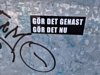 A black sticker with white text on a weathered surface. The text is in Swedish and translates to "Do it immediately, do it now."