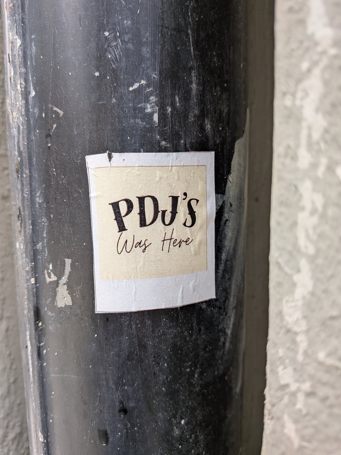 A roughly square sticker with a light beige or cream-colored central panel. It features a thin, dark brown inner border, followed by a wider white outer border. The text 'PDJ's' is prominently displayed in a bold, dark brown serif font, with 'Was Here' written below it in a smaller, elegant cursive script, also in dark brown. The sticker appears slightly worn, with minor creases and a small tear at the top edge, and is affixed to a dark, scratched, and textured pipe or pole.