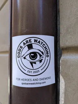 Street sticker A white sticker with a black logo that reads "GODS ARE WATCHING" in a circular arrangement around a stylized eye with a lightning bolt inside. Below the eye, it reads "EST. 2023" and "FOR HEROES AND DAEMONS". At the bottom of the sticker, the website "godsarewatching.com" is printed.