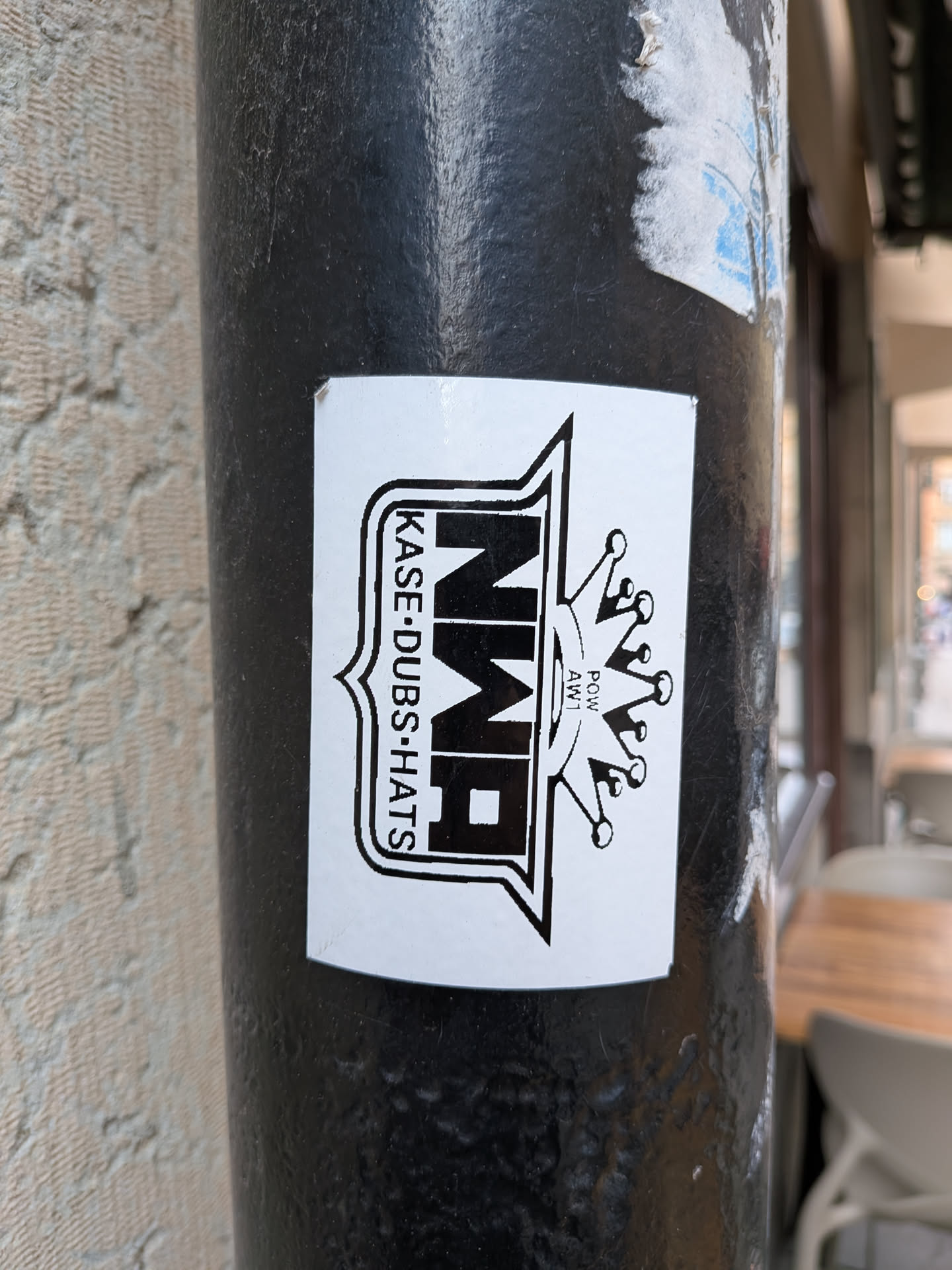 A rectangular white sticker featuring a central black and white design. The main element is a shield-like emblem with a thick black outline. Inside the shield, the letters 'NWR' are prominently displayed in a bold, blocky, stylized black font. To the left of 'NWR', the text 'KASE-DUBS-HATS' is vertically aligned in a smaller, sans-serif font. To the right of the 'NWR' and slightly outside the shield, a stylized black crown with five points, each topped with a small circle, is depicted. Below the crown, there is small text 'POW AW1'. The overall appearance is clean, graphic, and monochrome, suggesting a brand or crew identity.