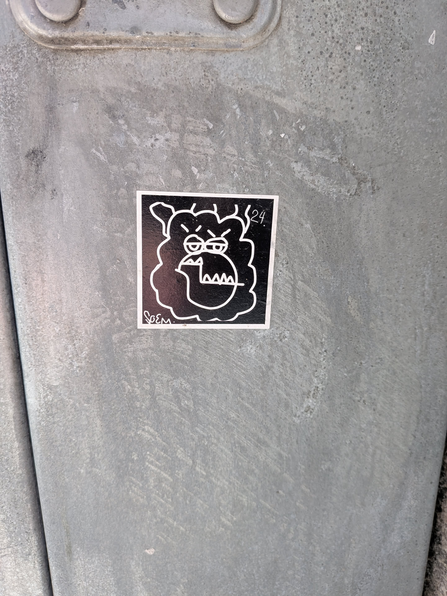A square sticker featuring a stylized, cartoonish character's head in white outline against a solid black background. The character has a thick, wavy outline suggesting fur or hair, prominent furrowed eyebrows, small oval eyes, and a large, open mouth revealing sharp, triangular teeth. Its expression conveys a sense of grumpiness or annoyance. In the bottom left corner, the signature 'Soem' is written in a stylized script, and in the top right corner, the numbers '24.' are visible.