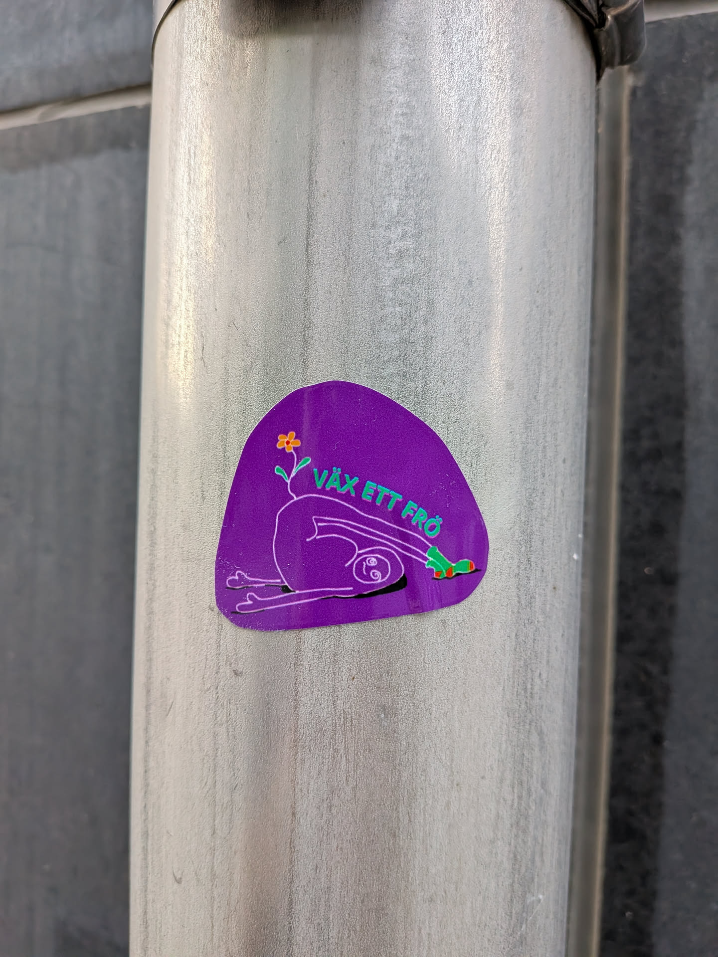 An organic-shaped purple sticker depicting a white-outlined figure in a deep stretch. A small orange flower sprouts from its back, and it wears green socks. Green Swedish text 'VÄX ETT FRÖ' curves above the figure.