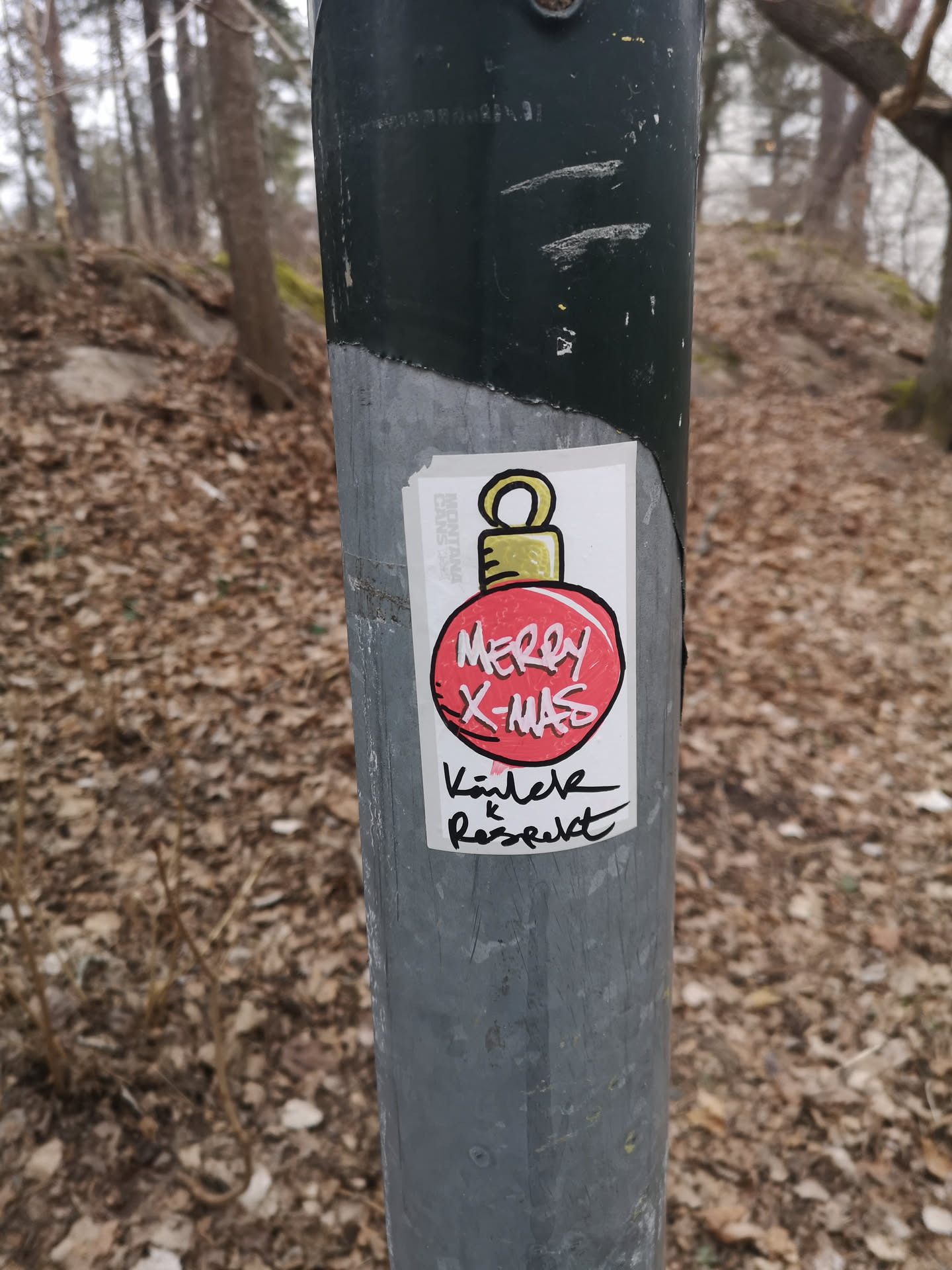 A hand-drawn style sticker featuring a red Christmas ornament with a golden top. The ornament has 'MERRY X-MAS' written in white, stylized capital letters. Below the ornament, in black handwritten script, are the words 'Kärlek & Respekt'. The sticker is rectangular with a white background, and a faint 'MONTANA CANS' logo is visible vertically along its left edge. It is affixed to a grey metal pole.