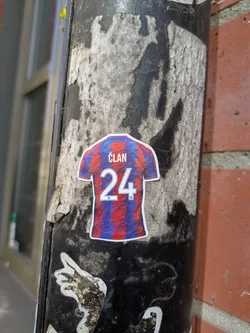 Street sticker This is a sticker featuring a soccer jersey with a modern design, showcasing a blend of red and blue hues. The name "ČLAN" is printed across the upper back, just above the number "24". The sticker is placed on a weathered black pole.