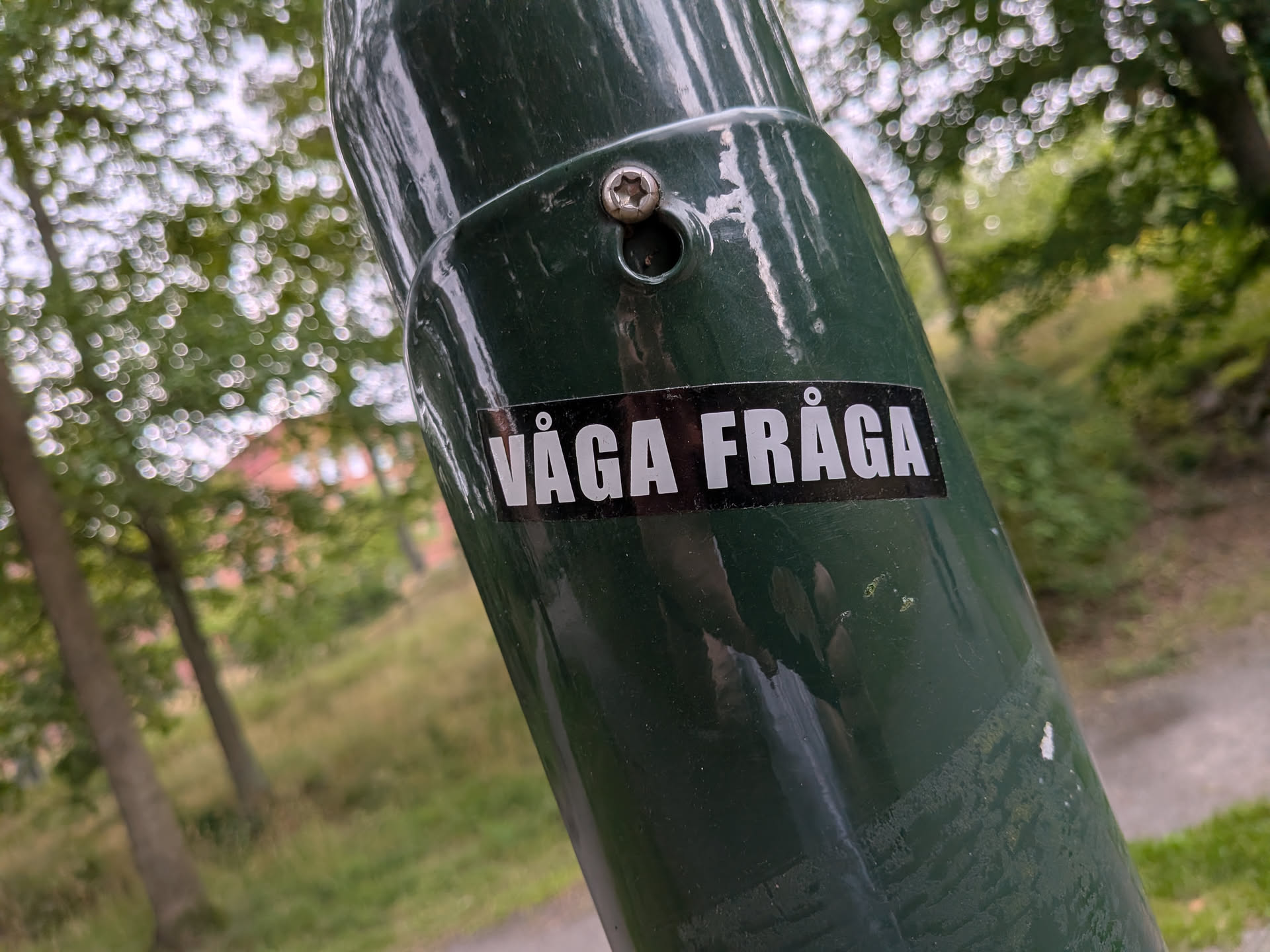 A simple rectangular black sticker with bold white sans-serif text reading 'VÅGA FRÅGA'. It is stuck to a dark green metal pole in an outdoor park setting.