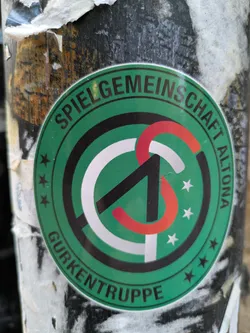 Street sticker Round green sticker with a logo and text. The logo is composed of a stylized letter 'S' in black, white and red. The text around the logo reads 'Spielgemeinschaft Altona Gurkentruppe'.