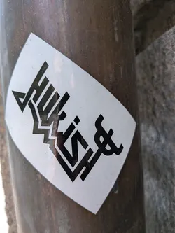 Street sticker The sticker is white with a bold, black graphic design. The design appears to be text or a stylized logo with sharp angles and a somewhat blocky appearance. The letters are thick and outlined with negative space, giving them a layered effect. The overall design has an urban or street-art aesthetic. At the far right of the design, there is a symbol that resembles a stylized trident or a fork.