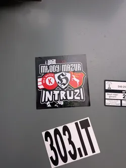 Street sticker The sticker is square-shaped with a black background. At the top, 'MŁODY MAZUR' is written in a distressed white, blocky font. Below this, there are two prominent emblems. On the left, a red circle with 'MKS MAZUR' written around its perimeter and a large white 'K' in the center, flanked by white and red stripes. On the right, a red shield features a white silhouette of a deer or stag, with 'ORLĘTA' written above it. The bottom of the sticker prominently displays 'INTRUZI' in a similar distressed white, blocky font, with some white splatter-like patterns around the text and edges.