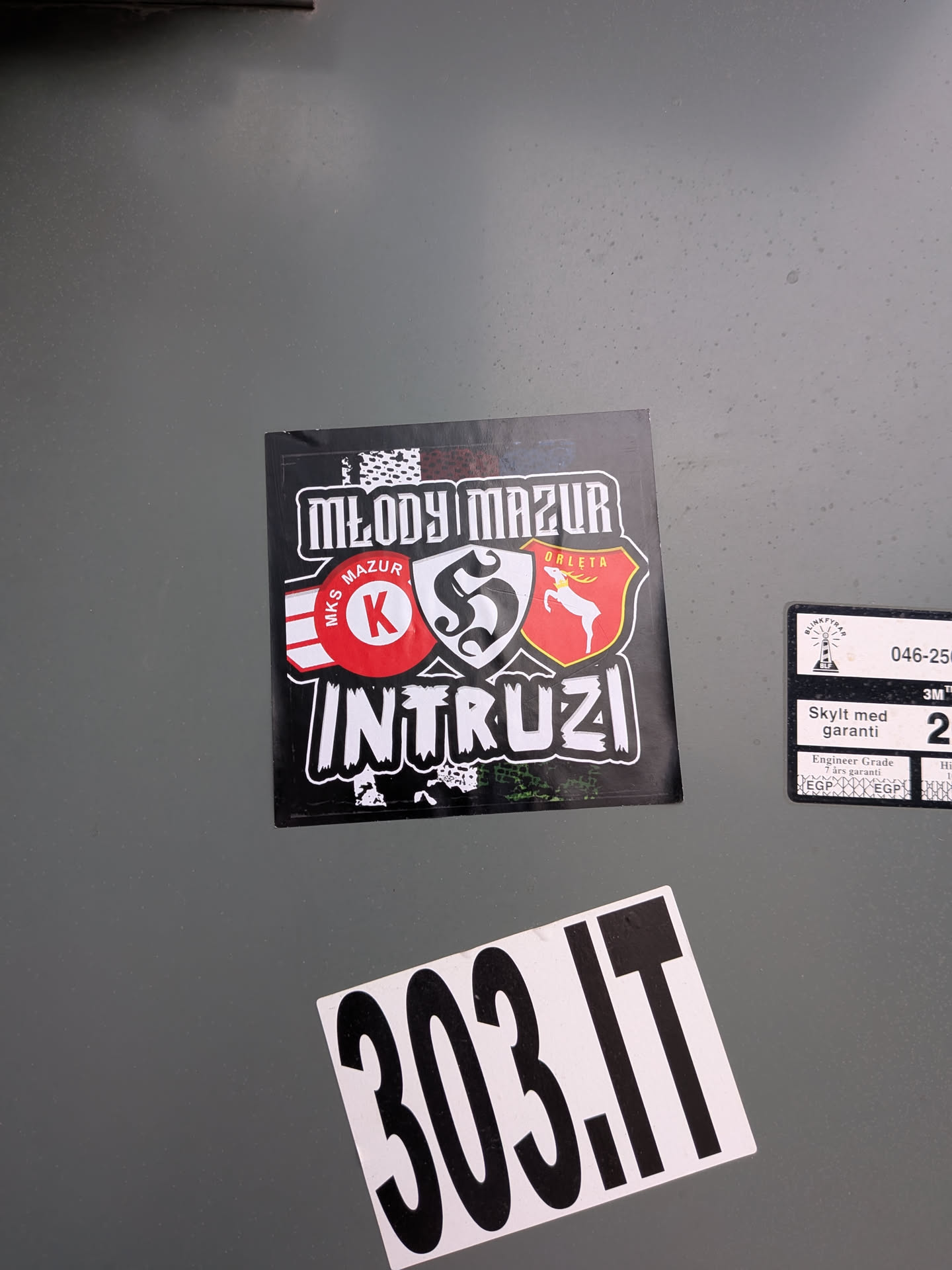 The sticker is square-shaped with a black background. At the top, 'MŁODY MAZUR' is written in a distressed white, blocky font. Below this, there are two prominent emblems. On the left, a red circle with 'MKS MAZUR' written around its perimeter and a large white 'K' in the center, flanked by white and red stripes. On the right, a red shield features a white silhouette of a deer or stag, with 'ORLĘTA' written above it. The bottom of the sticker prominently displays 'INTRUZI' in a similar distressed white, blocky font, with some white splatter-like patterns around the text and edges.