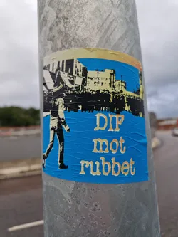 Street sticker A blue sticker with a black and white illustration of a person walking in front of a crowd and buildings. The text 'DIF mot rubbet' is written in a serif font below the illustration. The sticker is affixed to a metal pole.