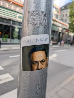 Street sticker The sticker displays a cropped portrait of Benjamin Netanyahu, showing his face from the forehead to just below the nose, with a serious or stern expression. His hair is dark, and his skin tone appears somewhat desaturated or aged, set against a dark, possibly green-tinted background. Above the portrait, on a white rectangular border, the phrase 'ISRAEL = NAZIS' is handwritten in blue capital letters. The sticker has slightly rounded corners and is affixed to a textured, metallic pole.