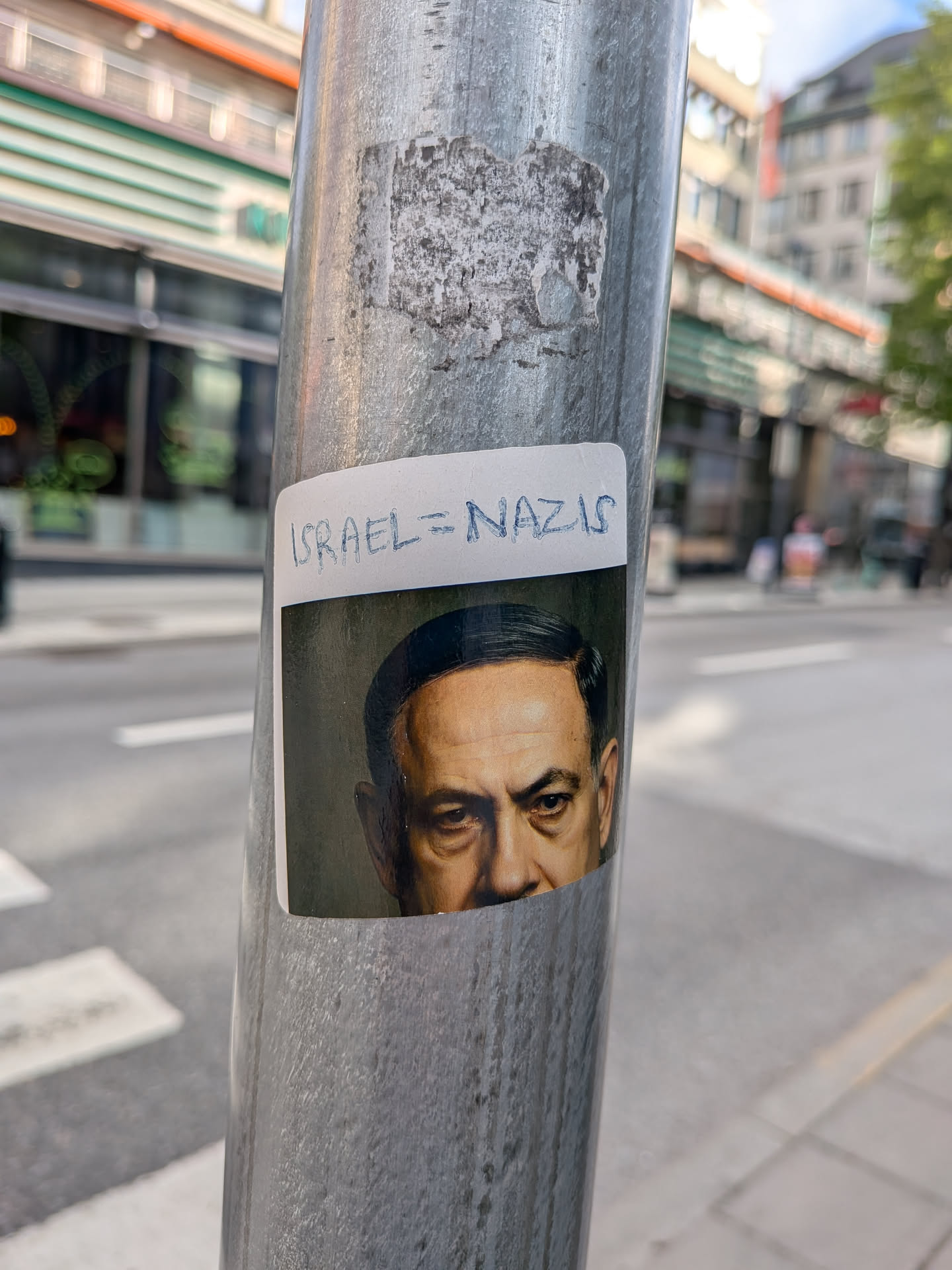 The sticker displays a cropped portrait of Benjamin Netanyahu, showing his face from the forehead to just below the nose, with a serious or stern expression. His hair is dark, and his skin tone appears somewhat desaturated or aged, set against a dark, possibly green-tinted background. Above the portrait, on a white rectangular border, the phrase 'ISRAEL = NAZIS' is handwritten in blue capital letters. The sticker has slightly rounded corners and is affixed to a textured, metallic pole.
