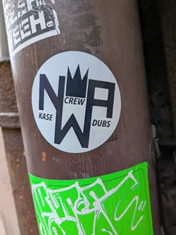 Street sticker A circular sticker with a light grey background, prominently featuring large, bold, black capital letters 'NWA'. The 'W' is stylized to form a crown-like peak at its center, connecting the 'N' and 'A'. Smaller, black sans-serif text reads 'KASE' to the left of the 'N', 'CREW' above the 'W', and 'DUBS' to the right of the 'A'. The overall design is clean, graphic, and monochromatic.