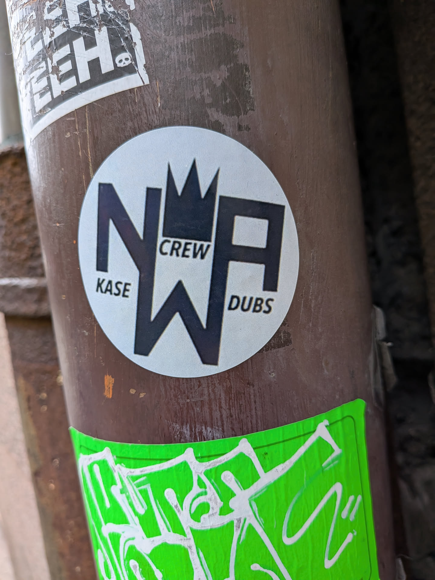 A circular sticker with a light grey background, prominently featuring large, bold, black capital letters 'NWA'. The 'W' is stylized to form a crown-like peak at its center, connecting the 'N' and 'A'. Smaller, black sans-serif text reads 'KASE' to the left of the 'N', 'CREW' above the 'W', and 'DUBS' to the right of the 'A'. The overall design is clean, graphic, and monochromatic.