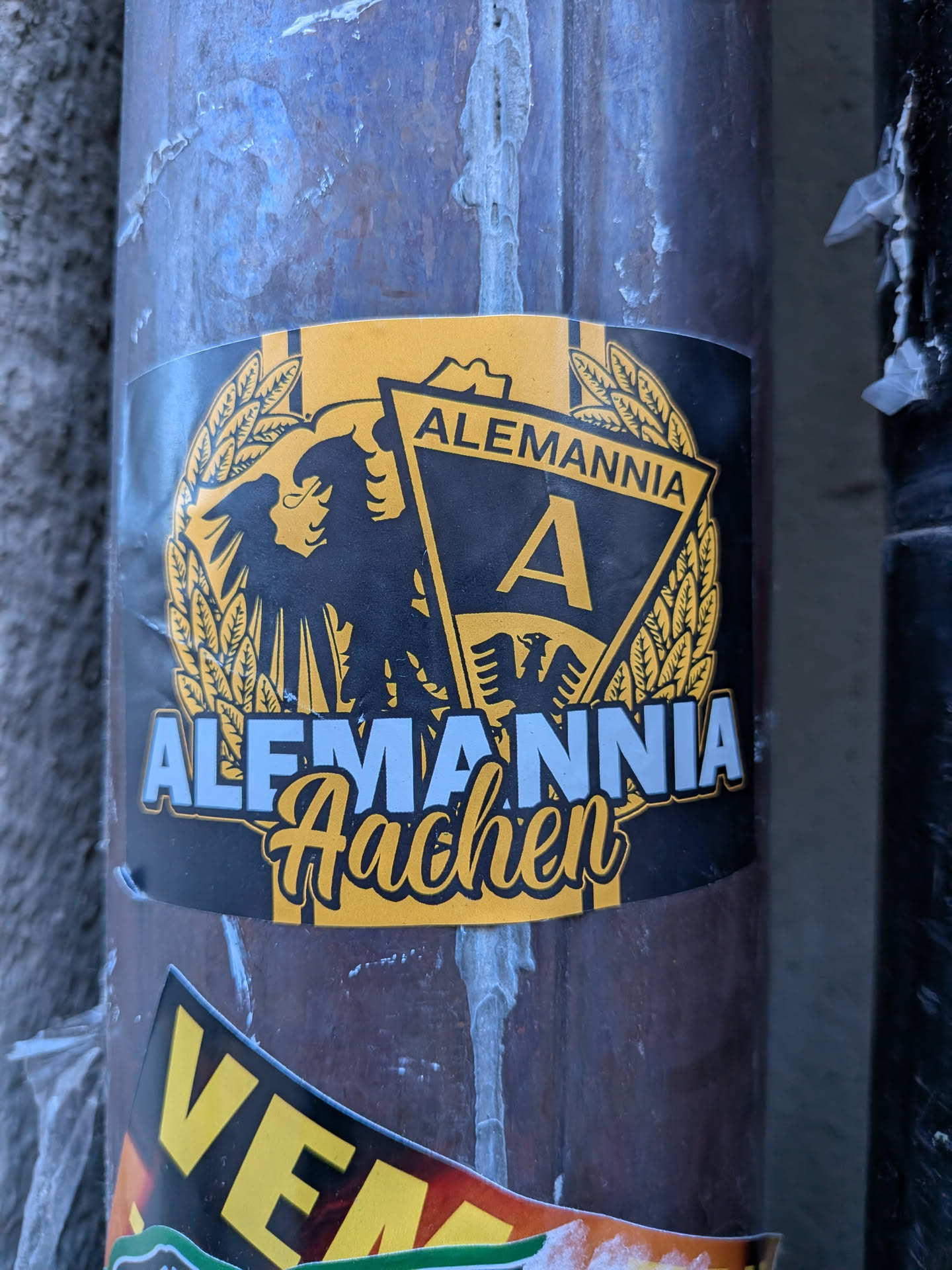 The sticker showcases the emblem of Alemannia Aachen, a German football club. It is predominantly black and yellow, with some light blue/white text. The central design features a large black eagle with spread wings, facing left, set against a yellow wreath-like background. To the right of the eagle, there's a black triangular pennant with a yellow border, containing a large yellow capital 'A'. Above this pennant, the word 'ALEMANNIA' is written in yellow capital letters on a black banner. Below the main crest, 'ALEMANNIA' is written in prominent light blue/white block letters with a yellow outline, and beneath that, 'Aachen' is written in a flowing yellow script font with a black outline. The sticker has a slightly worn appearance, indicating it's a physical sticker on a pole.