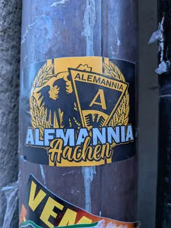 Street sticker The sticker showcases the emblem of Alemannia Aachen, a German football club. It is predominantly black and yellow, with some light blue/white text. The central design features a large black eagle with spread wings, facing left, set against a yellow wreath-like background. To the right of the eagle, there's a black triangular pennant with a yellow border, containing a large yellow capital 'A'. Above this pennant, the word 'ALEMANNIA' is written in yellow capital letters on a black banner. Below the main crest, 'ALEMANNIA' is written in prominent light blue/white block letters with a yellow outline, and beneath that, 'Aachen' is written in a flowing yellow script font with a black outline. The sticker has a slightly worn appearance, indicating it's a physical sticker on a pole.
