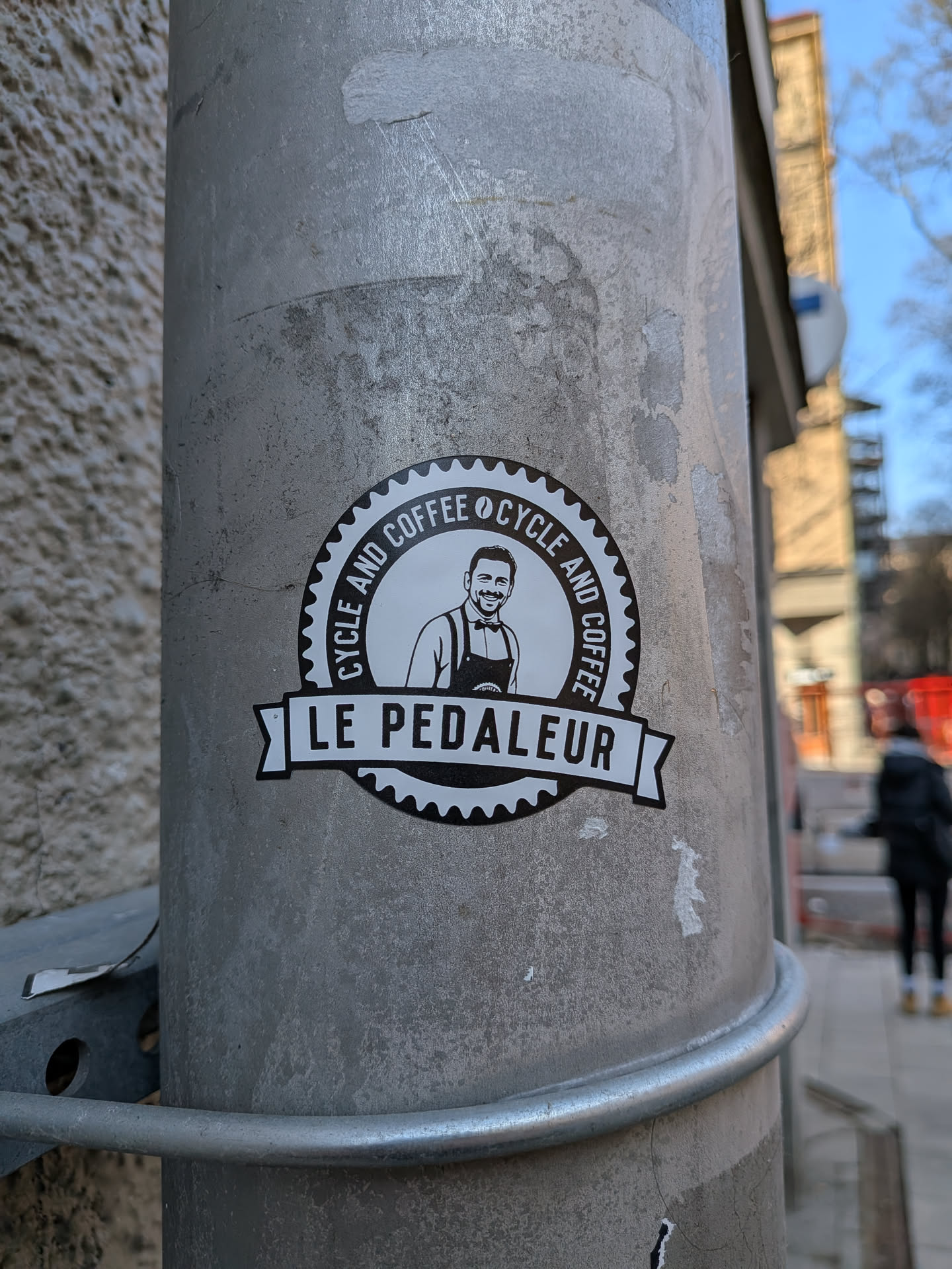 A circular, black and white sticker featuring a smiling man with a beard and short dark hair, wearing a collared shirt and an apron. The outer edge of the circle is designed like a bicycle gear or cog. Around the central illustration, the text 'CYCLE AND COFFEE' is repeated. Below the circle, a black banner with pointed ends displays the text 'LE PEDALEUR' in white capital letters. The overall design is clean, minimalist, and logo-like.