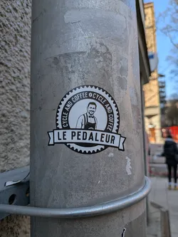 Street sticker A circular, black and white sticker featuring a smiling man with a beard and short dark hair, wearing a collared shirt and an apron. The outer edge of the circle is designed like a bicycle gear or cog. Around the central illustration, the text 'CYCLE AND COFFEE' is repeated. Below the circle, a black banner with pointed ends displays the text 'LE PEDALEUR' in white capital letters. The overall design is clean, minimalist, and logo-like.