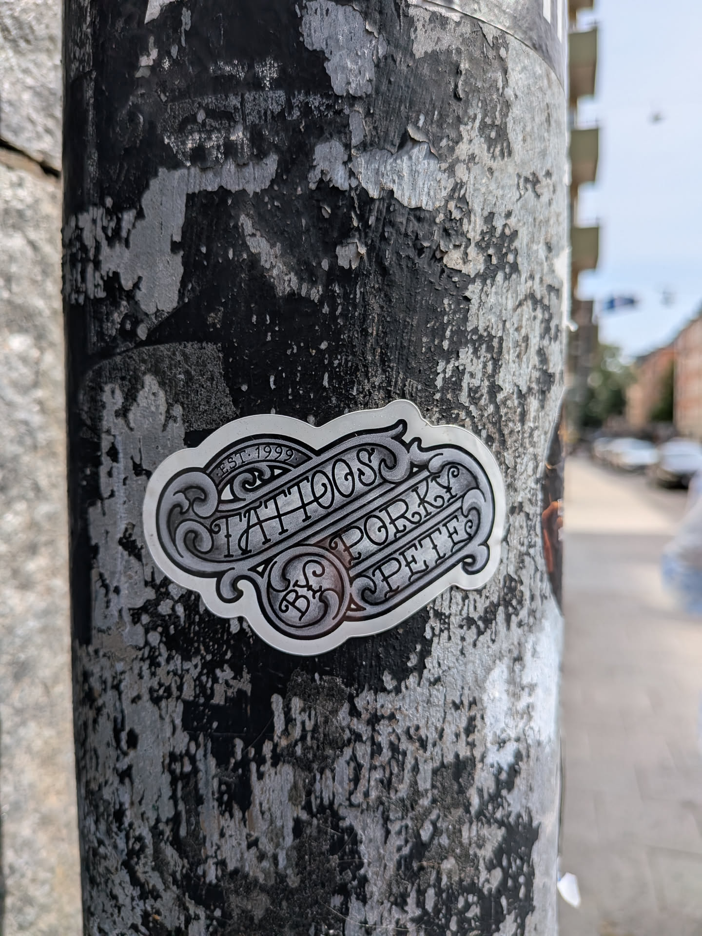 A die-cut, metallic-grey sticker featuring intricate, vintage-style filigree and scrollwork. The stylized black lettering reads 'EST. 1999 TATTOOS BY PORKY PETE'. It is adhered to a heavily weathered, textured pole.