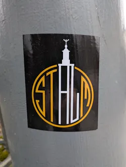 Street sticker The sticker is a black rectangular shape with slightly rounded corners, adhered to a textured grey pole. Its central design features a white, stylized architectural structure resembling a tower or spire, topped with a small cross-like or winged element. This tower is integrated with a prominent circular outline in a vibrant yellow-gold color. Within this circle, the word 'STALIA' is formed. The 'S' and 'T' are rendered in yellow-gold, while the 'ALIA' part is in white, with the vertical stroke of the 'L' extending upwards to form a significant portion of the tower structure. The overall aesthetic is clean, modern, and logo-like, utilizing a limited palette of black, white, and yellow-gold.