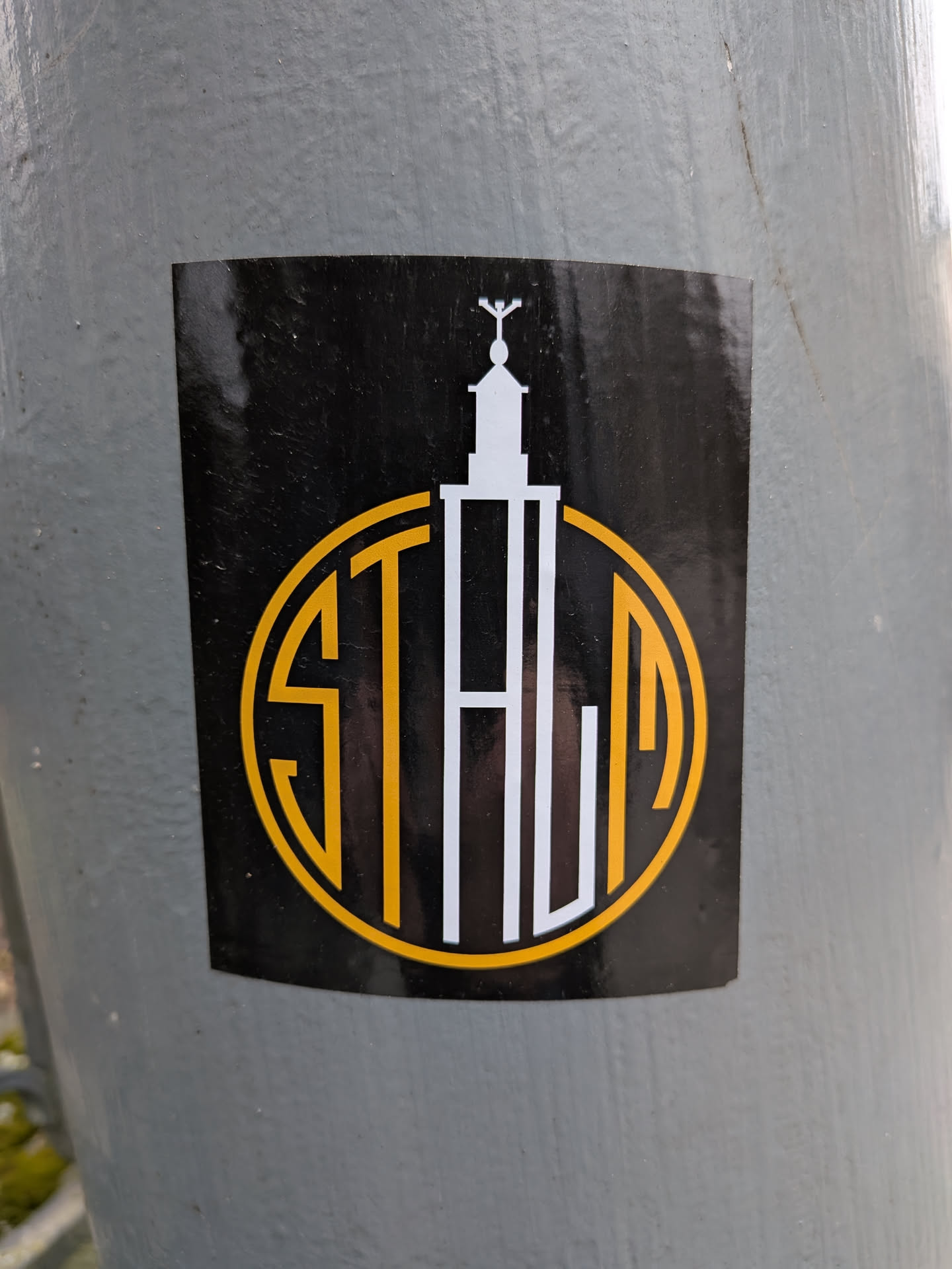 The sticker is a black rectangular shape with slightly rounded corners, adhered to a textured grey pole. Its central design features a white, stylized architectural structure resembling a tower or spire, topped with a small cross-like or winged element. This tower is integrated with a prominent circular outline in a vibrant yellow-gold color. Within this circle, the word 'STALIA' is formed. The 'S' and 'T' are rendered in yellow-gold, while the 'ALIA' part is in white, with the vertical stroke of the 'L' extending upwards to form a significant portion of the tower structure. The overall aesthetic is clean, modern, and logo-like, utilizing a limited palette of black, white, and yellow-gold.