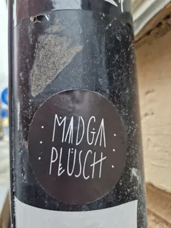 A dark-colored round sticker with the text ": MADGA : PLÜSCH :" written in a stylized font. The sticker is affixed to a dark pole, possibly outdoors. 
