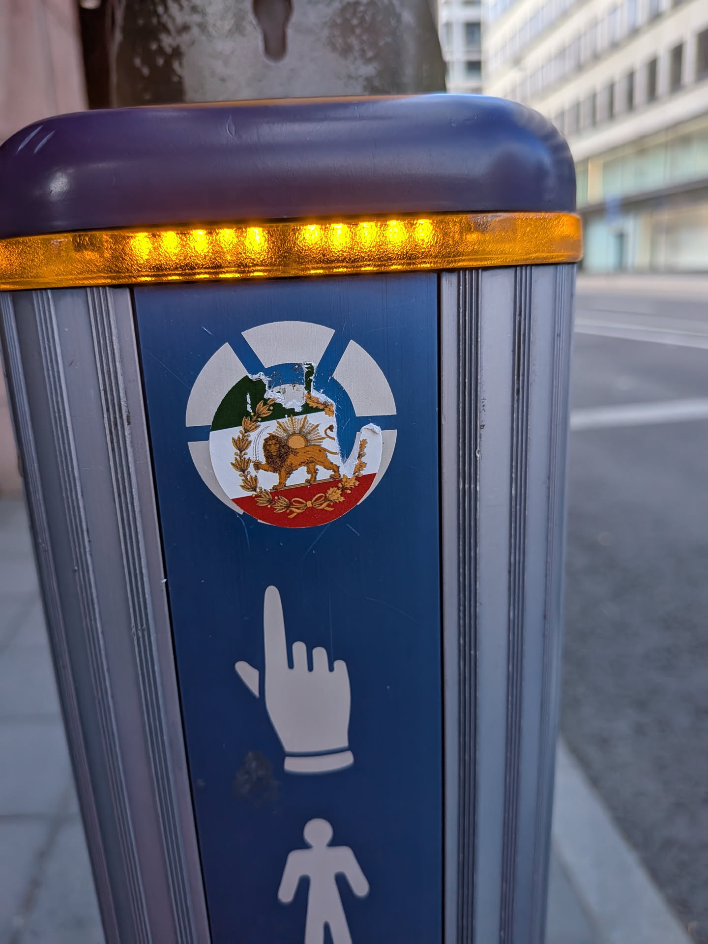 A circular, worn sticker depicting the historical Iranian Lion and Sun emblem over a green, white, and red tricolor background, pasted on a blue pedestrian signal post.