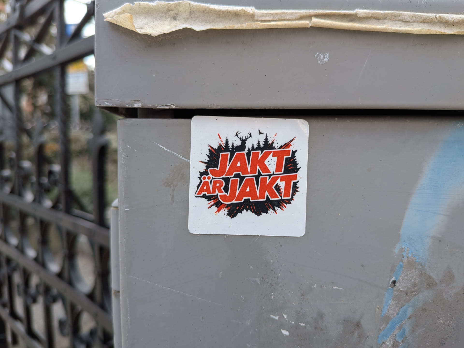 Square white sticker featuring bold red text "JAKT ÄR JAKT" over a black splash background containing silhouettes of a stag, pine trees, and a bird.