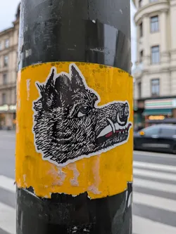 Street sticker The sticker features a black and white illustration of a wolf's head in profile. The wolf has sharp teeth bared, a red tongue visible, and a piercing blue eye. The fur is depicted with intricate black lines. The sticker has a white border and is affixed to a yellow surface with a hexagonal reflective pattern. The edges of the sticker appear slightly worn.