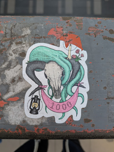 Street sticker This sticker features a ram skull with turquoise tentacles emerging from it. A pink banner is draped beneath, with the number "1000" printed on it. A lit lantern hangs from one side of the banner, and roses and a candle are on the other side.