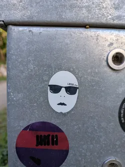 Street sticker An oval white sticker featuring a simple face graphic with black sunglasses, two small dots for nostrils, a stylized mouth, and the text 'LBSB.nu' printed above the right lens.
