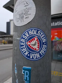 The sticker is circular with a prominent blue border. Inside the border, the text 'KIELER S.V. HOLSTEIN' is written in white capital letters, arranged in a semi-circle. Below this, 'VON 1900' is also in white capital letters. The center of the sticker features a shield with a red and white jagged pattern around the edges. Within this, a white emblem with black lines depicts a stylized boat or crescent shape, possibly representing a historical symbol of the region or club. The overall design is clean and emblematic, typical of sports club insignia.
