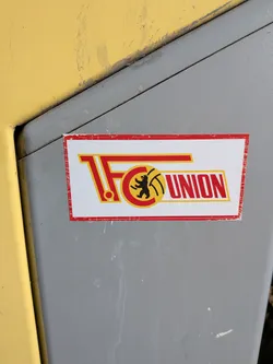 Street sticker A rectangular sticker featuring the logo of 1. FC Union Berlin. The sticker has a white background with a distressed red border. The main design consists of large, stylized red letters '1. FC' with yellow outlines on the left. Integrated into the 'C' is a circular emblem with a black bear holding a yellow football, all within a red and yellow outline. To the right, the word 'UNION' is written in bold red letters with a yellow outline. The overall color scheme is red, yellow, black, and white. The sticker shows signs of wear, particularly around its edges.