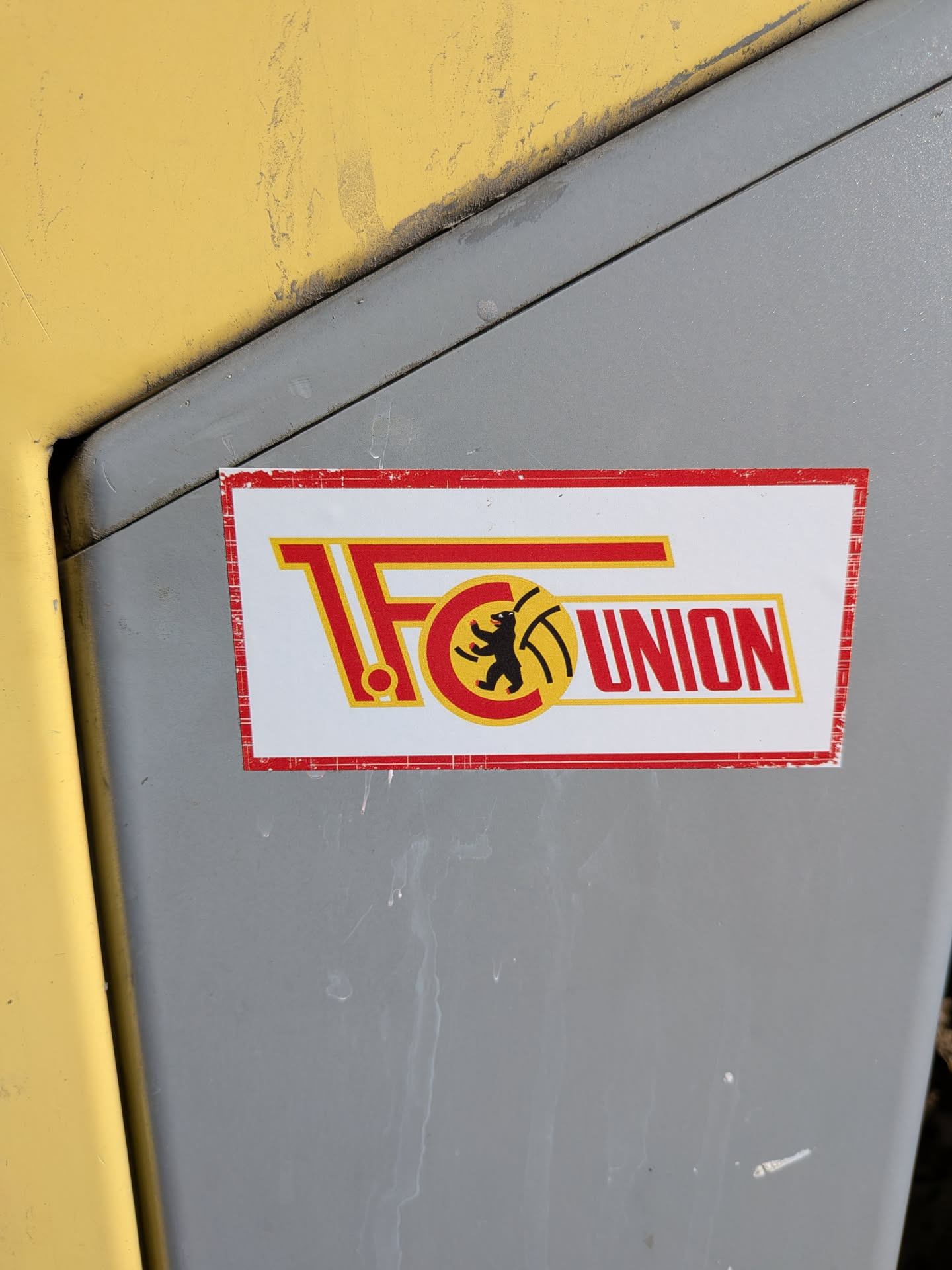 A rectangular sticker featuring the logo of 1. FC Union Berlin. The sticker has a white background with a distressed red border. The main design consists of large, stylized red letters '1. FC' with yellow outlines on the left. Integrated into the 'C' is a circular emblem with a black bear holding a yellow football, all within a red and yellow outline. To the right, the word 'UNION' is written in bold red letters with a yellow outline. The overall color scheme is red, yellow, black, and white. The sticker shows signs of wear, particularly around its edges.