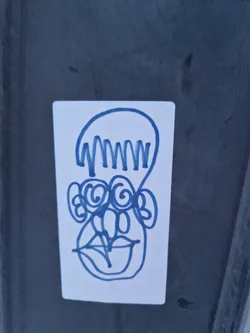Street sticker Blue ink drawing of a face with a unique hairstyle. The style is minimalistic and slightly abstract.  The sticker is affixed to a dark gray surface.