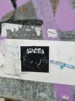 Black and white sticker with the word AGNOSIA in a circle and two hands underneath.  The text b.t.x boys is visible below the main sticker.