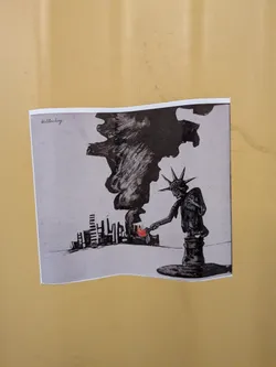 Street sticker A square sticker featuring a black-and-white ink illustration. It depicts the Statue of Liberty leaning down to set fire to a city skyline with her torch. A massive plume of black smoke billows upward. The flame is the only colored element, rendered in red. The name 'Hillersberg' is written in the top-left corner.