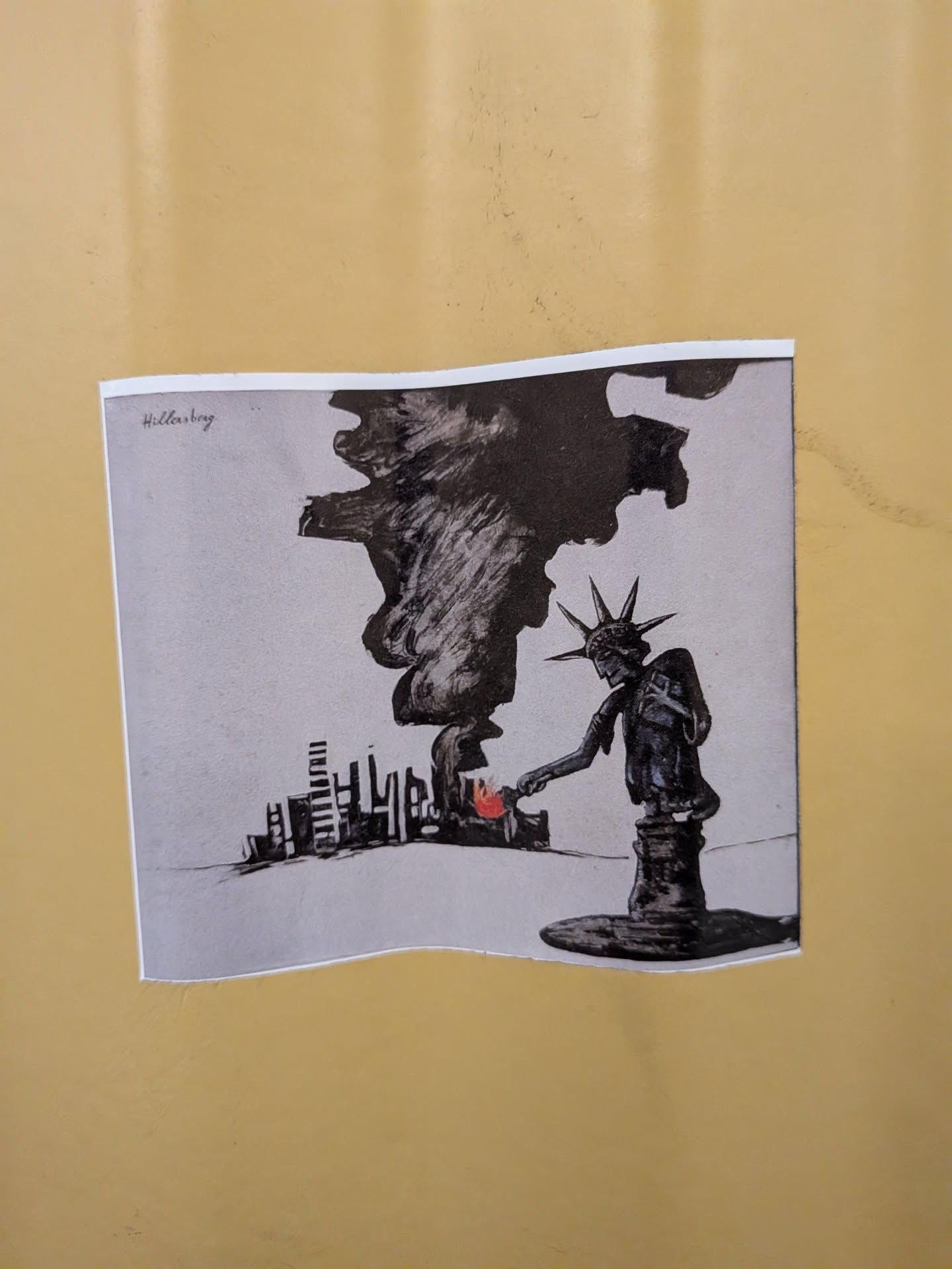 A square sticker featuring a black-and-white ink illustration. It depicts the Statue of Liberty leaning down to set fire to a city skyline with her torch. A massive plume of black smoke billows upward. The flame is the only colored element, rendered in red. The name 'Hillersberg' is written in the top-left corner.
