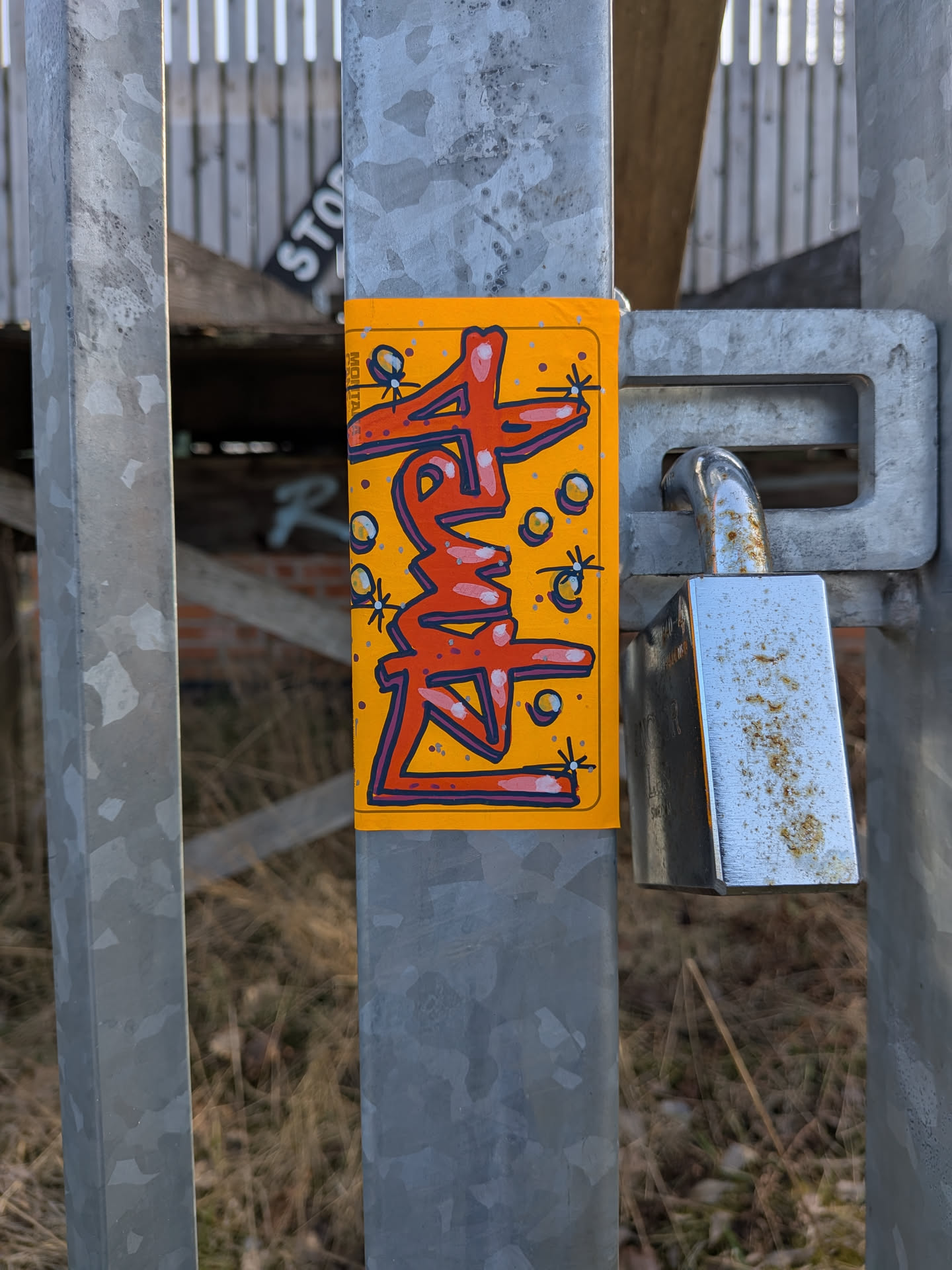 A rectangular sticker with a bright orange background adhered to a galvanized metal pole. The central design features highly stylized, graffiti-style lettering in a prominent red color with a dark blue outline and lighter blue/white highlights, giving it a dimensional look. The letters are abstract but appear to form a word, possibly 'FREAK' or similar. Surrounding the lettering are small, light yellow or gold circular 'bubble' elements and tiny 'sparkle' effects, depicted as small crosses with dots at their ends. Along the left edge of the sticker, vertically oriented, the word 'MONTANA' is visible in small, dark text. The sticker has slightly rounded corners.