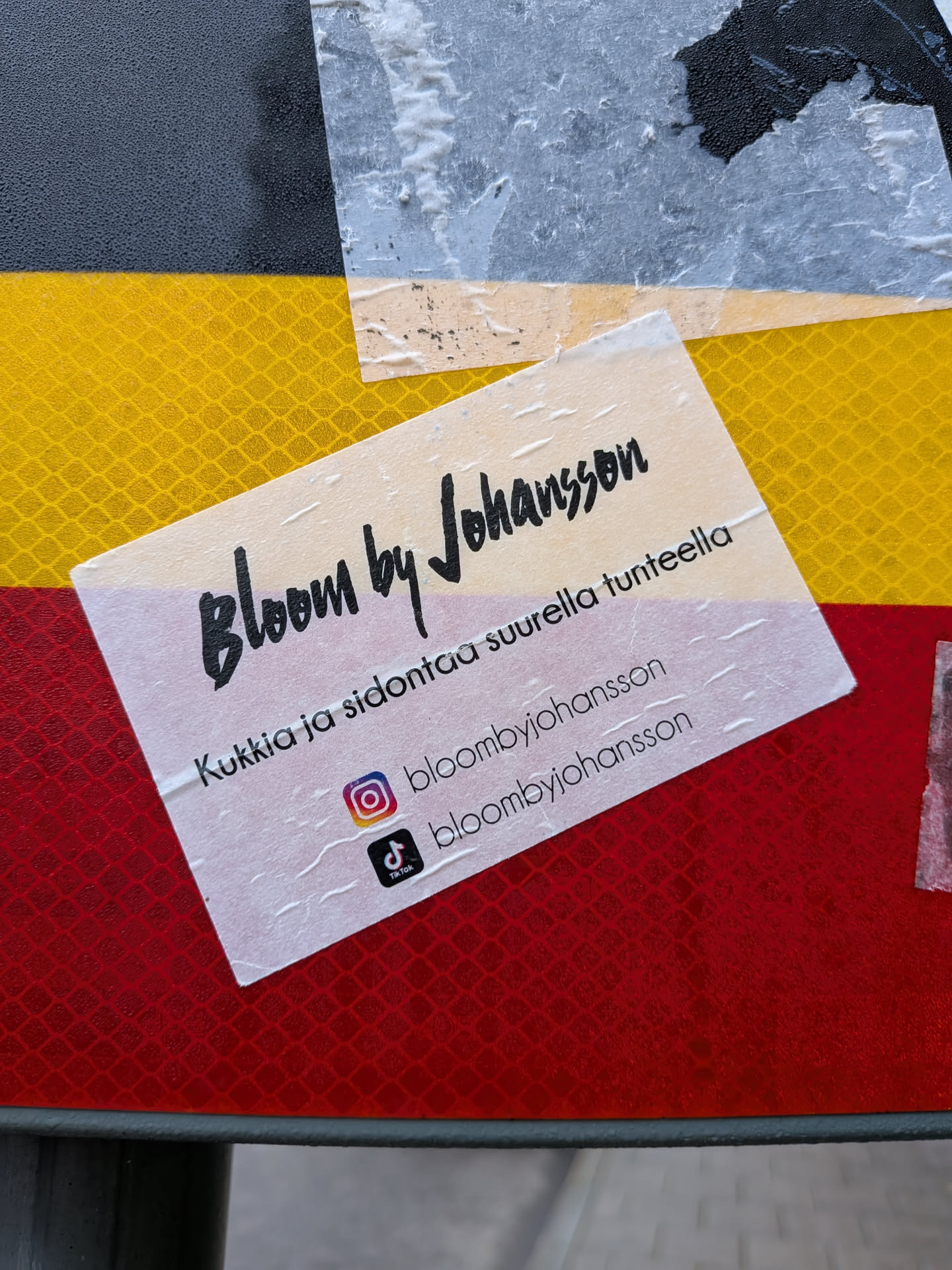 Street sticker A semi-transparent sticker with a white background and black text. The top line reads 'Bloom by Johansson' in a stylish, calligraphic font. Below this, in a smaller, standard font, is the text 'Kukkia ja sidontaa suurella tunteella'. The sticker also features social media icons: an Instagram logo followed by '@bloombyjohansson', and a TikTok logo followed by '@bloombyjohansson'. The sticker is placed over a background of alternating yellow and red reflective diamond patterns.