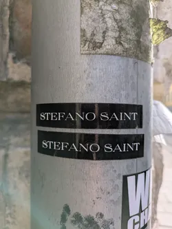 Street sticker The image features a sticker adhered to a metallic pole. The sticker has a black background with the text "STEFANO SAINT" printed in white. There are two identical stickers placed one above the other. Below those there is another sticker that partially appears in the frame
