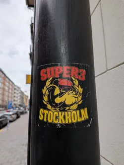 Square black sticker featuring a cartoon bulldog in a red cap smoking a cigar, framed by a yellow laurel wreath. 'SUPER3' is written in red at the top, and 'STOCKHOLM' in yellow at the bottom.