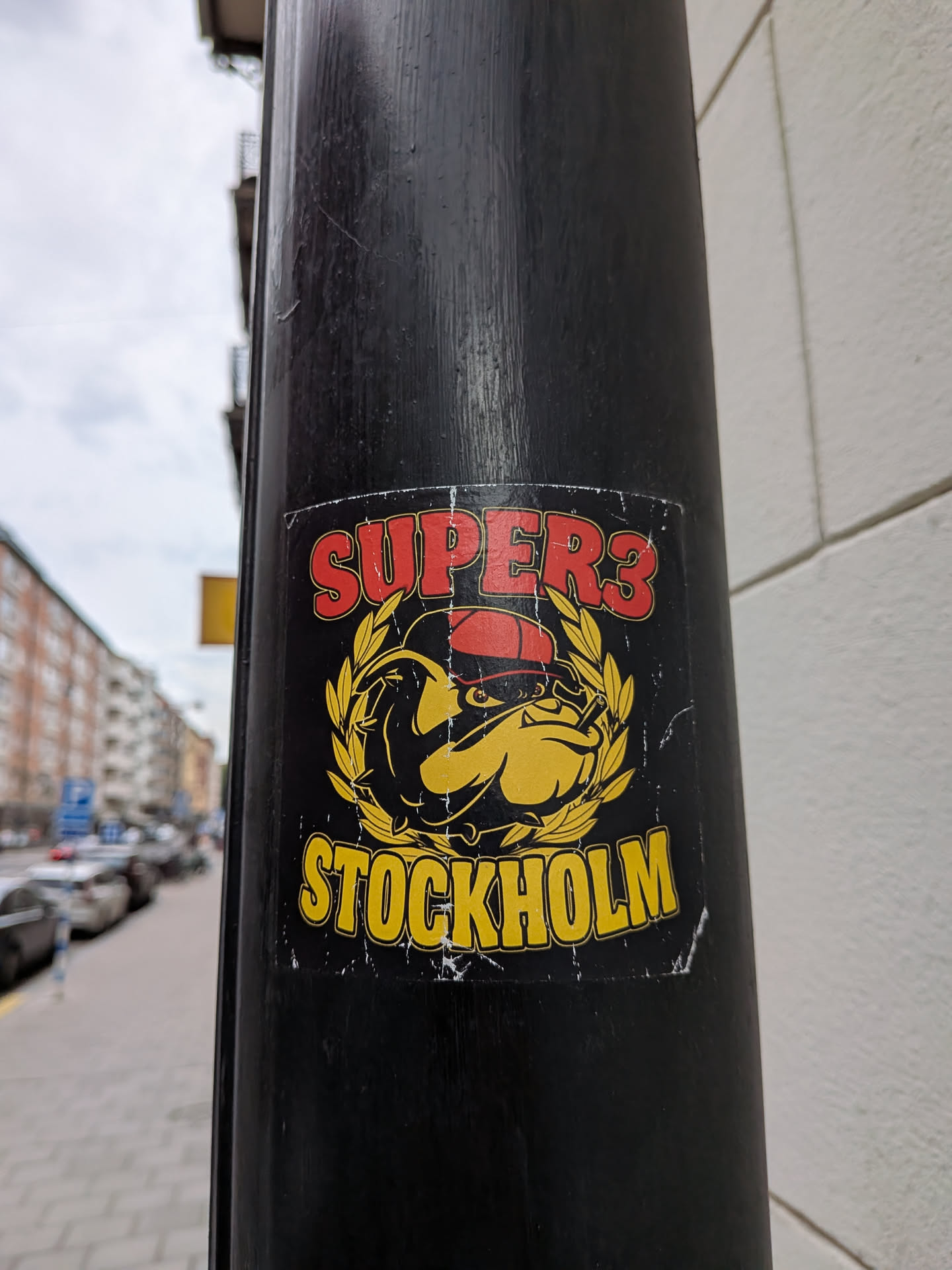 Square black sticker featuring a cartoon bulldog in a red cap smoking a cigar, framed by a yellow laurel wreath. 'SUPER3' is written in red at the top, and 'STOCKHOLM' in yellow at the bottom.