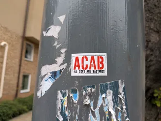 Street sticker The sticker is rectangular with a white background and a red border. Prominently displayed in large, red, sans-serif capital letters are the letters 'ACAB'. Below this, in smaller black sans-serif capital letters, is the text 'ALL COPS ARE BASTARDS'. The sticker appears to be slightly worn at the edges and is affixed to a grey, cylindrical pole, with other torn and weathered stickers surrounding it.