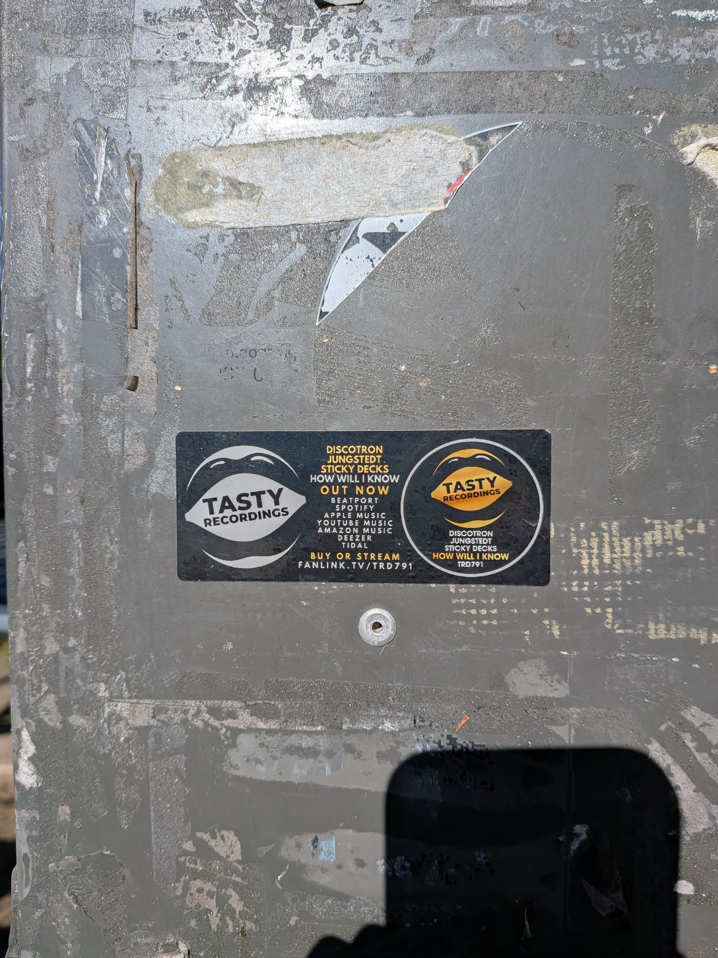 A black rectangular sticker featuring white and gold 'Tasty Recordings' mouth logos. The center contains text detailing a music release by Discotron, Jungstedt, and Sticky Decks, listing available streaming platforms and a fanlink URL.