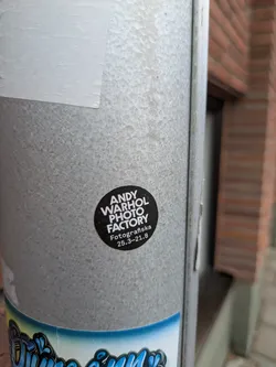 Street sticker The sticker is circular with a black background and white text. The main text, 'ANDY WARHOL PHOTO FACTORY', is prominently displayed in a bold, sans-serif font, stacked over three lines. Below this, in a slightly smaller font, are the words 'Fotografiska' and the dates '25.3-21.8', indicating an exhibition period. The design is minimalist and clean, focusing solely on the text content. The sticker is affixed to a textured, galvanized metal pole.