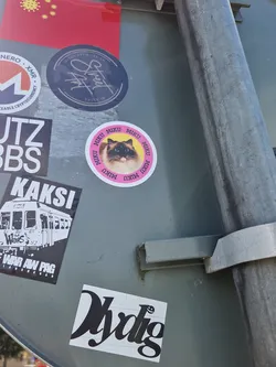 A round sticker featuring a photo of Miku, a fluffy Ragdoll cat. The cat's name is repeated around the image in a circle. The sticker is attached to a grey metal surface along with other stickers.