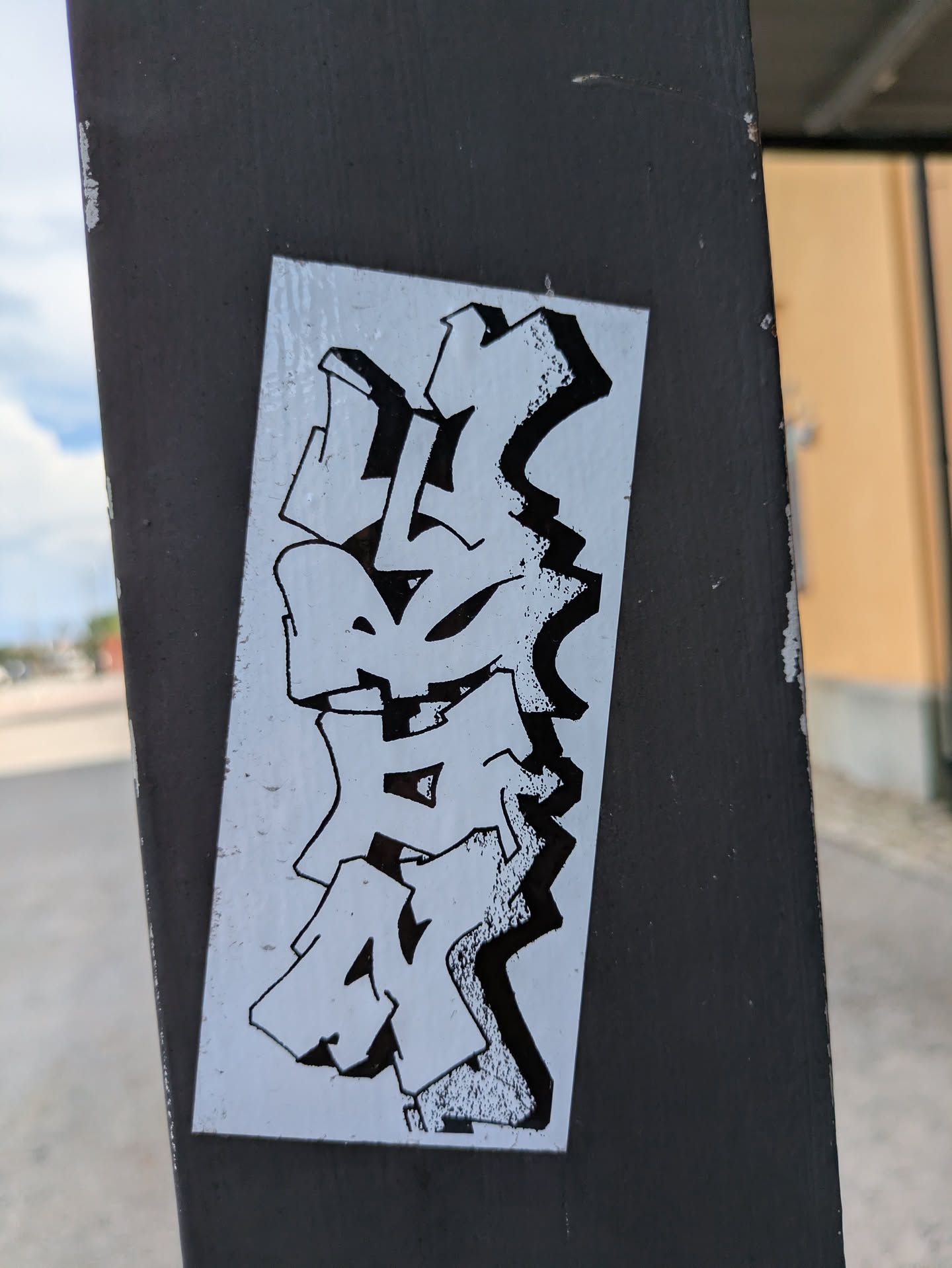 Street sticker A rectangular sticker featuring highly stylized, abstract graffiti lettering in black and white. The letters are stacked vertically, with thick black outlines and a textured, speckled fill that creates a gritty, shadowed effect, giving them a three-dimensional appearance. The background of the sticker is plain white. The overall style is characteristic of 'wildstyle' graffiti, making the specific text difficult to decipher. The sticker is affixed to a dark, textured metal pole.